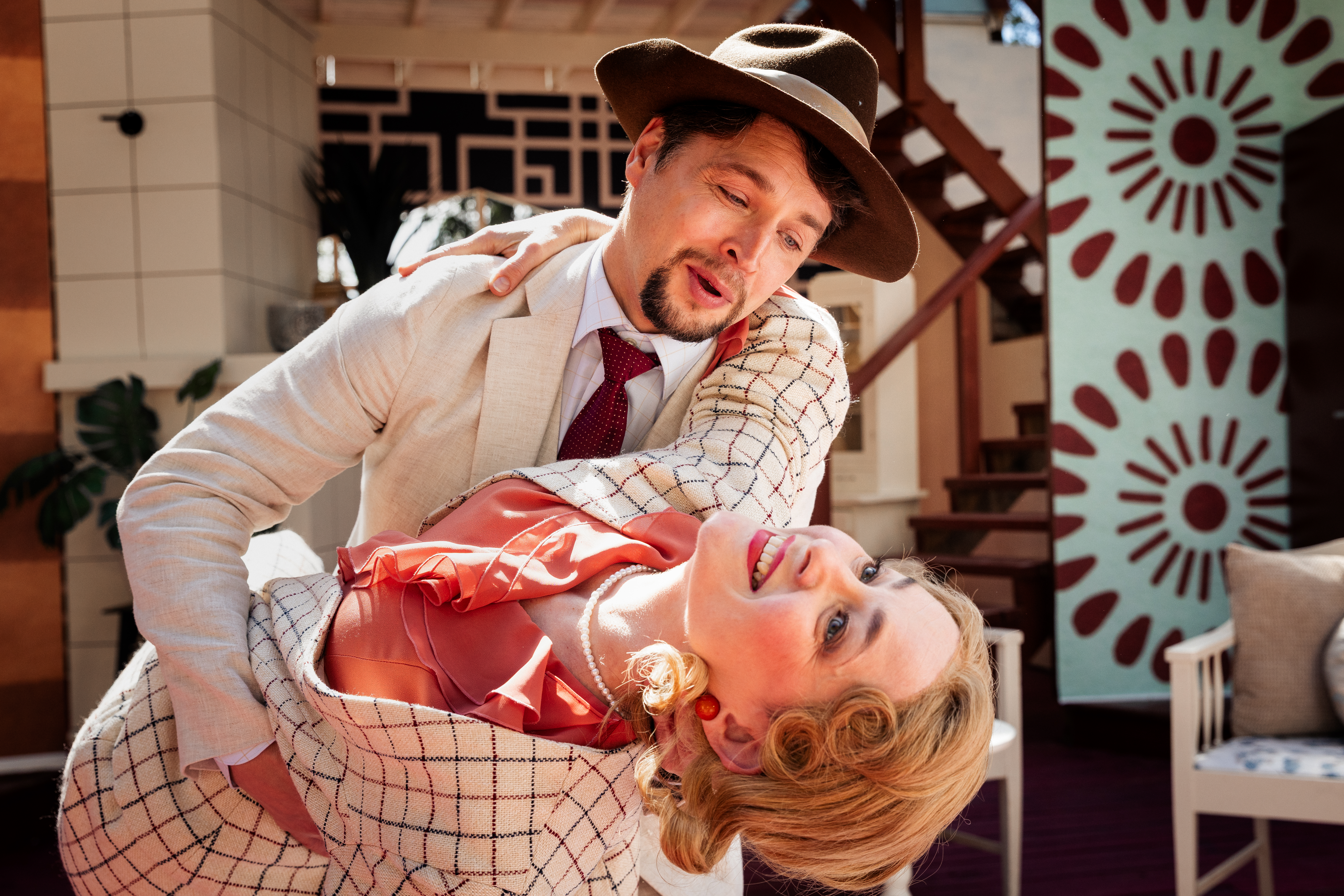 Kaksi näyttelijää teatterilavalla tanssiasennossa: hatullinen henkilö pitelee toista henkilöä taaksepäin taivutettuna. Molemmilla on vaaleat, 1900-luvun alun tyyliä muistuttavat asut, ja taustalla näkyy sisätilaa esittävä lavastus portaikkoineen ja kuvioituine seinäpintoineen kirkkaassa valaistuksessa.