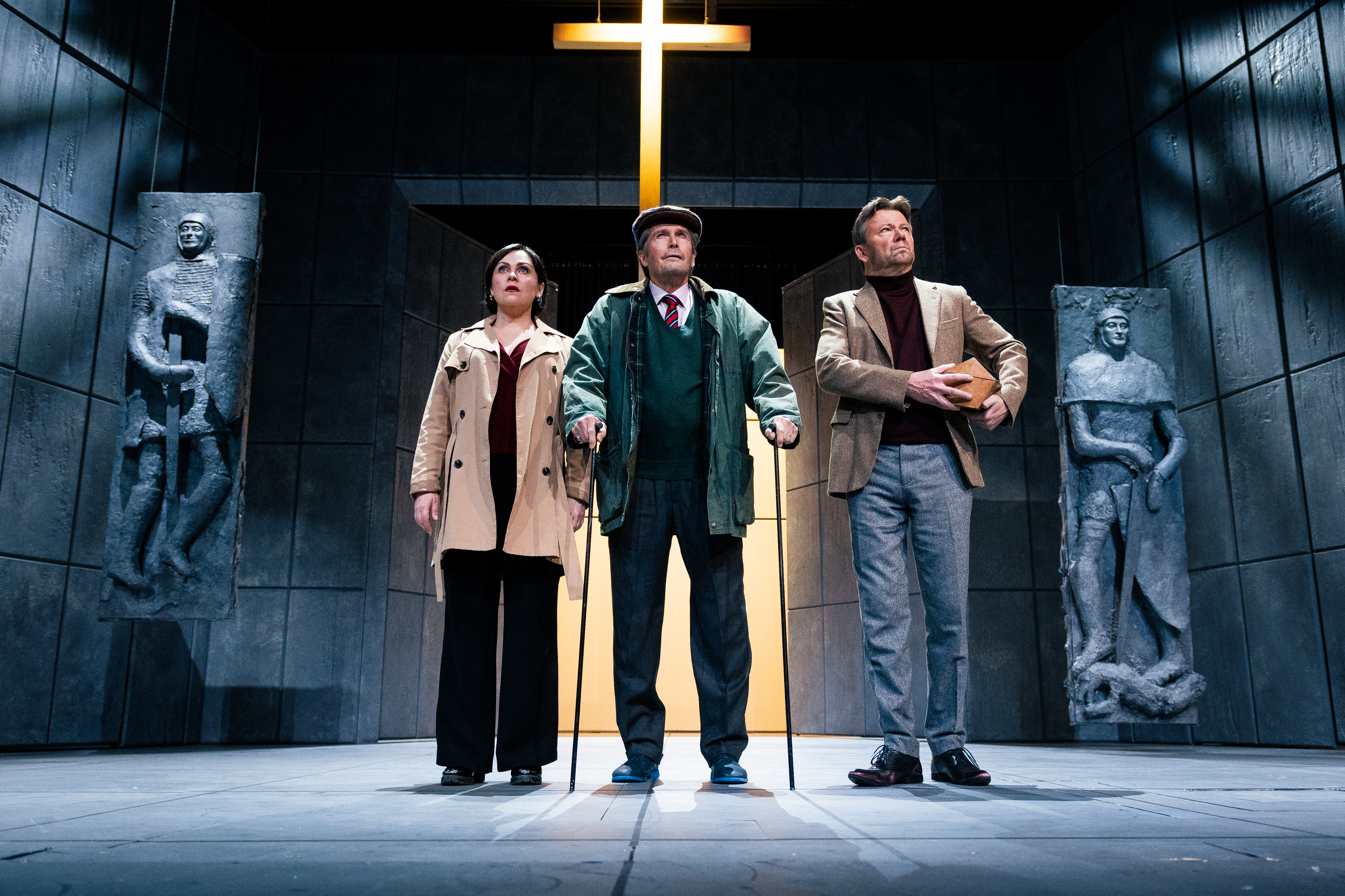 Teatteriesityksen näyttämökuva: kolme henkilöä seisoo rinnakkain tummassa, kivimäisessä tilassa. Taustalla loistaa suuri valaistu risti. Keskellä vanhempi mies nojaa kävelykeppiin, vasemmalla nainen vaaleassa takissa ja oikealla mies pitelee laatikkoa. Heitä ympäröivät seinäreliefeihin veistetyt ihmishahmot, ja tunnelma on vakava ja dramaattinen.