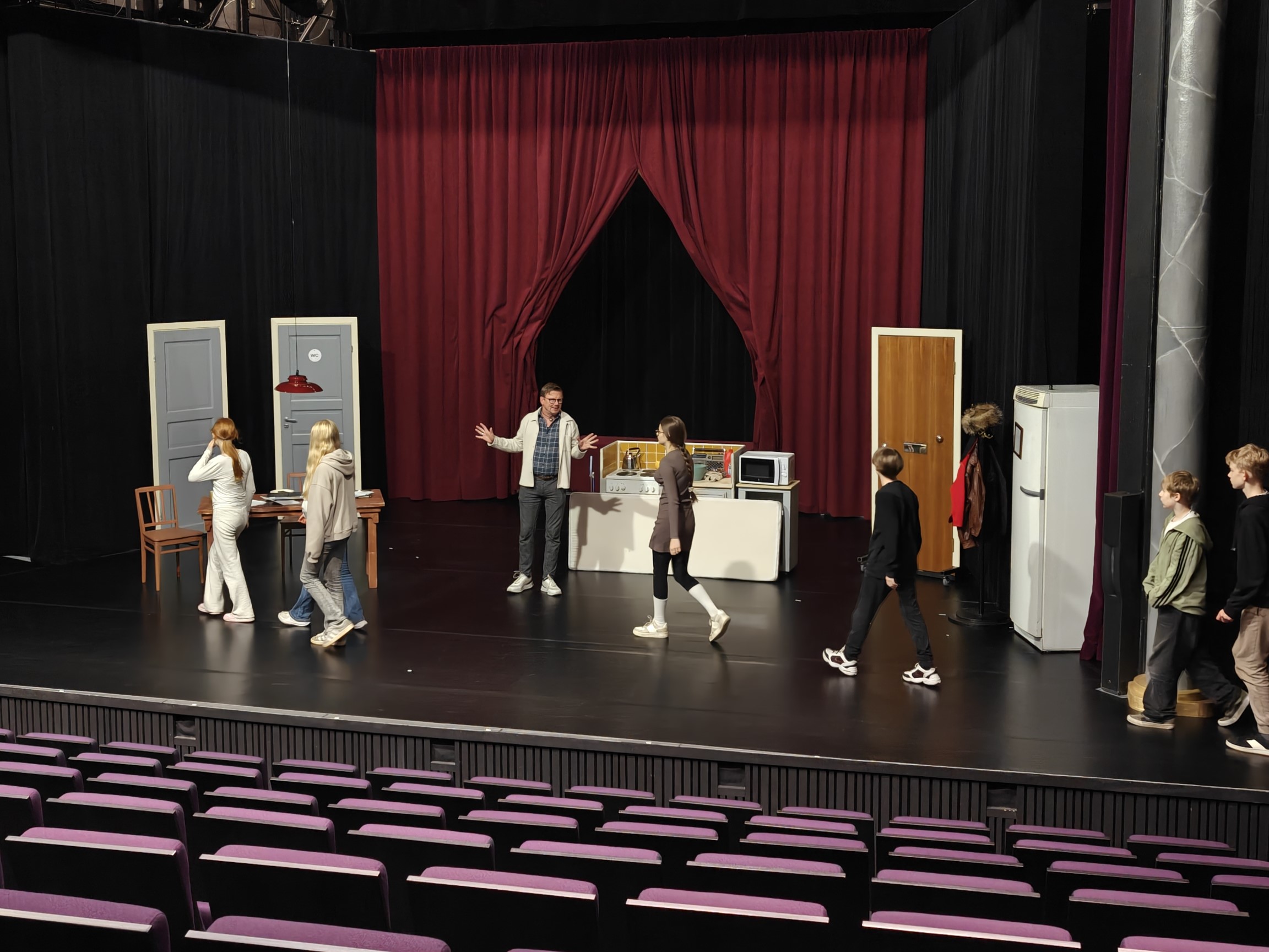 Ryhmä koululaisia tutustuu teatterin näyttämöön. Oppilaat kävelevät lavalla teatterin edustajan johdolla lavastetussa näyttämöympäristössä, jossa on pöytä, tuoleja, ovia, jääkaappi ja keittiötaso. Näyttämön taustalla näkyvät suuret punaiset verhot, ja etualalla on tyhjä katsomo violetteine istuimineen.