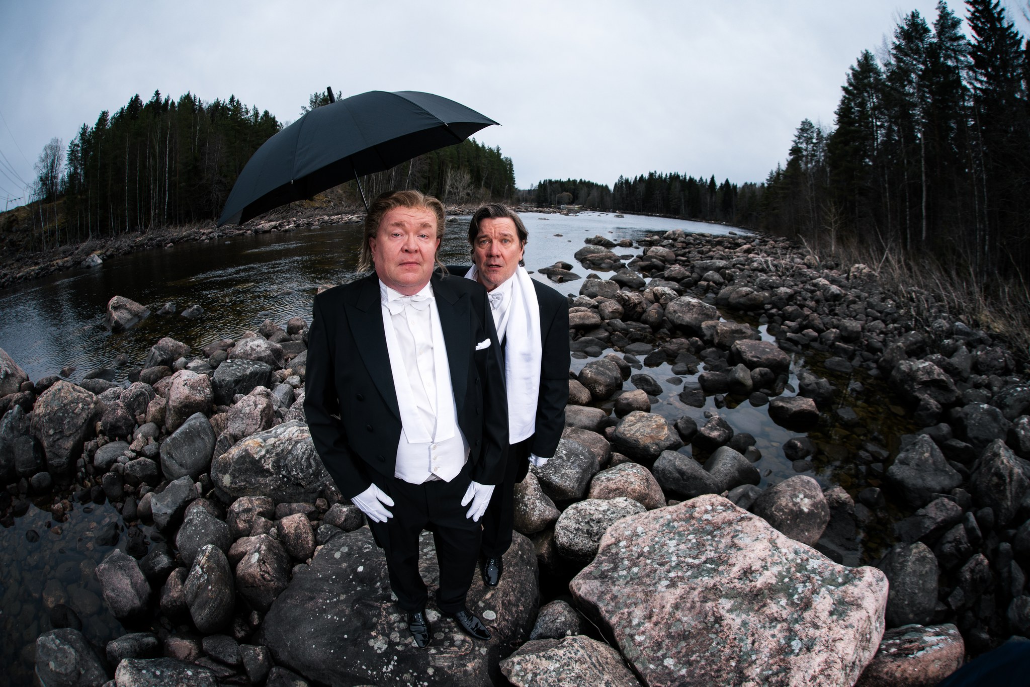 Kaksi frakkiin pukeutunutta henkilöä seisoo kivikkoisella joenrannalla. Toisen yläpuolella on avoin musta sateenvarjo. Taustalla näkyy virtaava joki, runsaasti kiviä sekä metsää harmaan pilvisen taivaan alla.