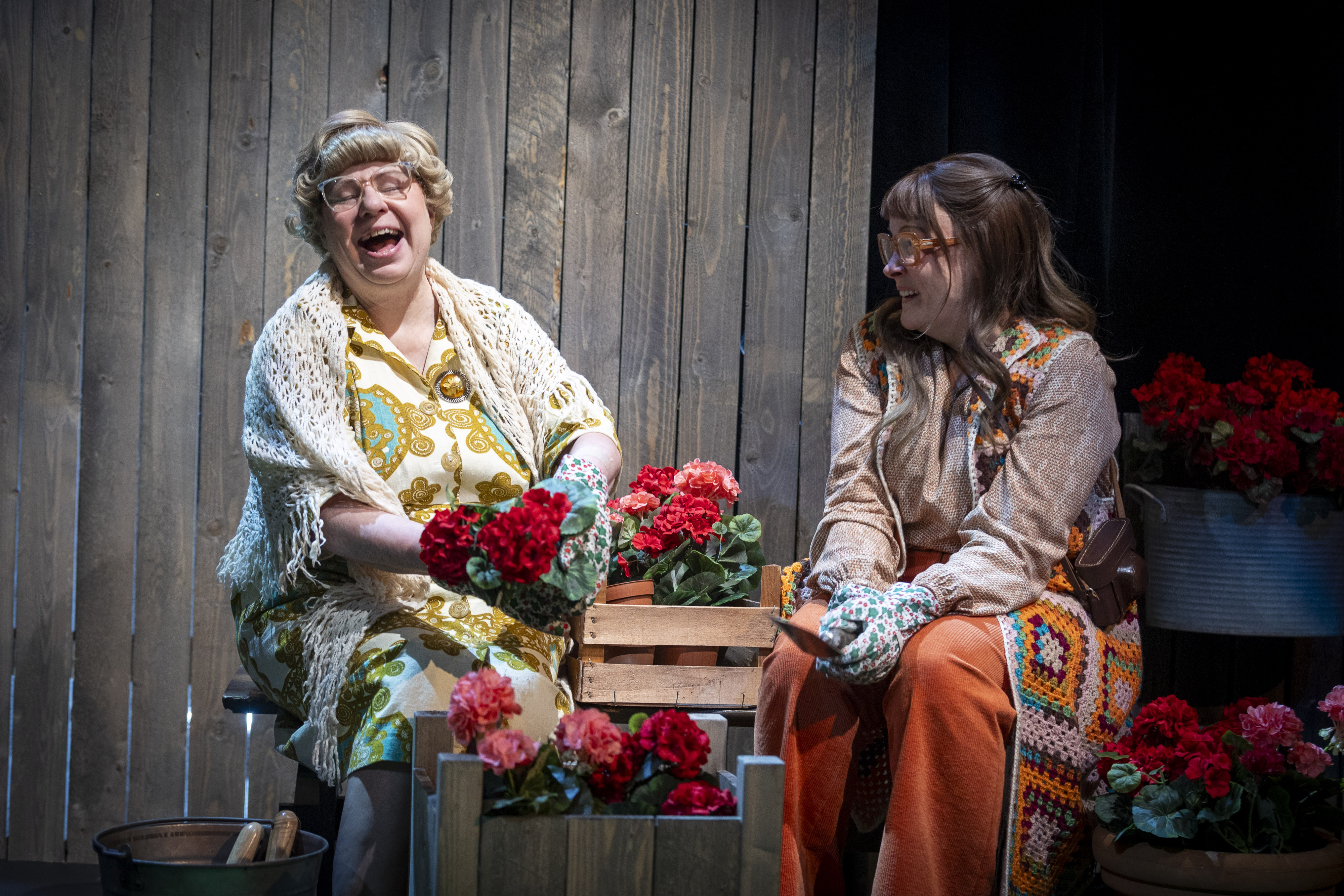 Kaksi nauravaa naishenkilöä istuu kukkalaatikoiden ääressä. Taustalla näkyy lautaseinä.