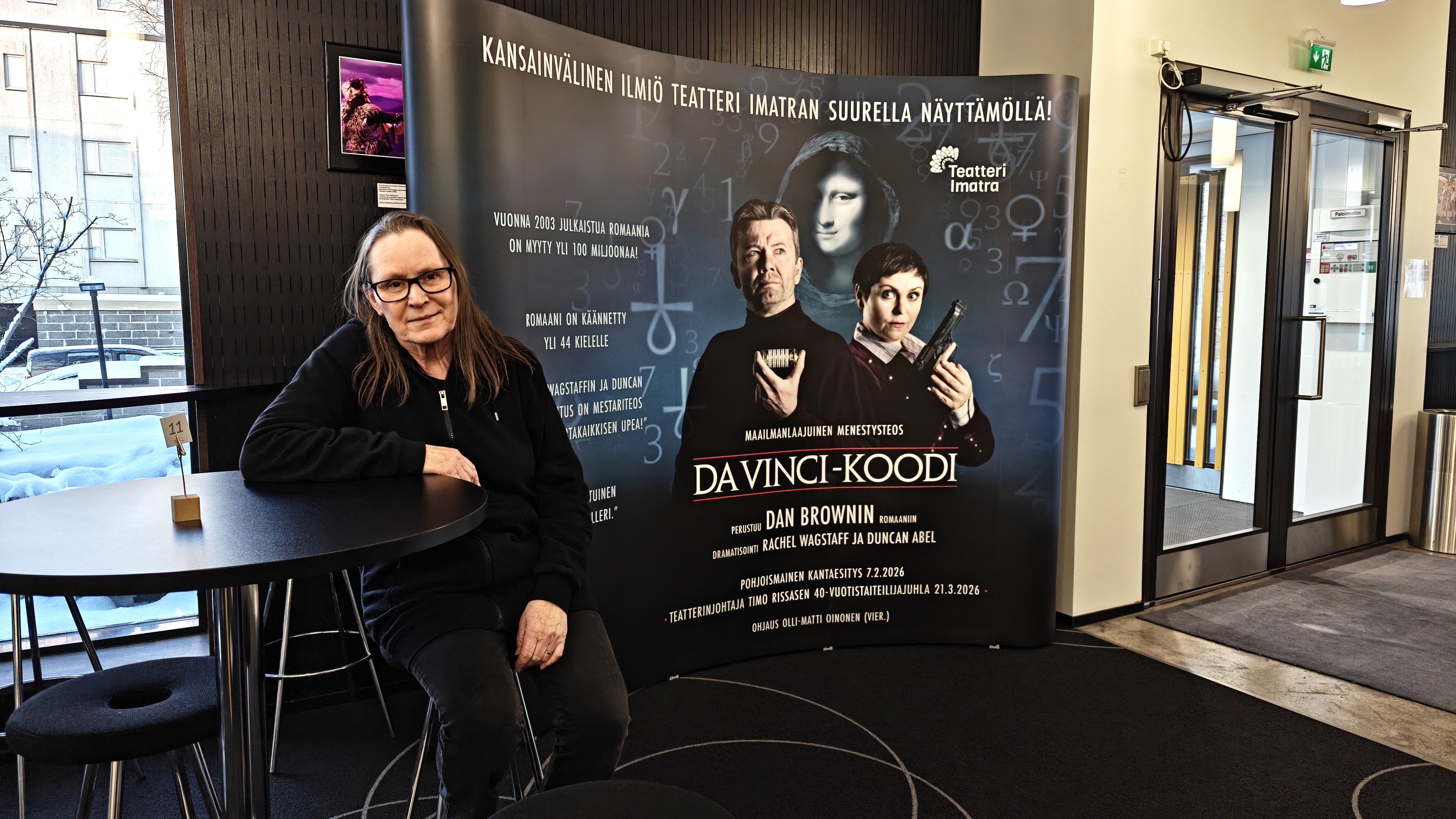 Näyttelijä istuu tuolilla ja nojaa pöytään. Taustalla näkyy ikkunat sekä Da Vinci -koodin mainosseinä.