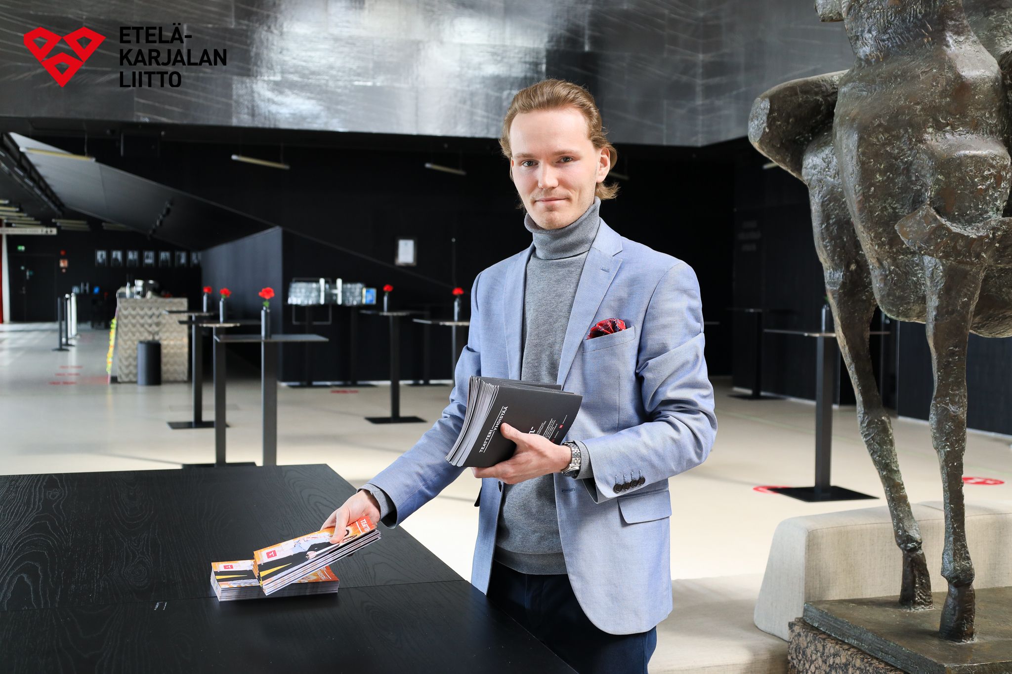 Henkilö (Miika Asikainen) pitelee käsissään teatterin esitteitä. Taustalla näkyy Lappeenrannan kaupunginteatterin lämpiö, jossa muun muassa pöytiä. Kuvan päällä Etelä-Karjalan liiton logo.