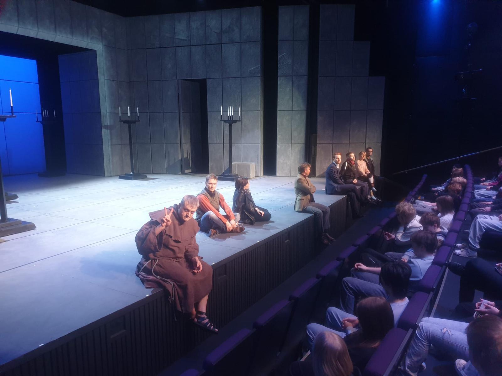 Teatterin näyttämö, jossa useita esiintyjiä istuu lattialla ja näyttämön reunalla. Taustalla on harmaa, kivimäinen lavastus ja kynttiläjalustoja. Etualalla näkyy katsomossa istuva yleisö, joka seuraa esitystä.